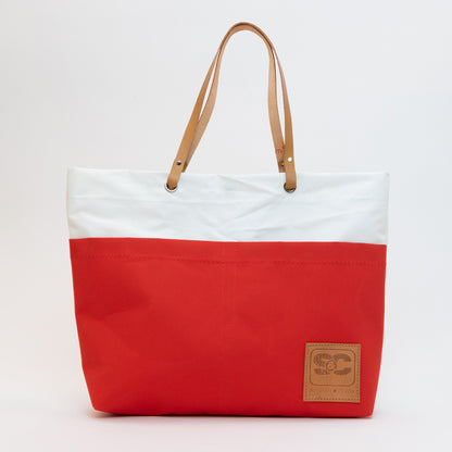 Recycled Sails Tote Bag. Large red tote with white panel at top connecting to leather handles through eyelets. Leather patch to right corner of red base.