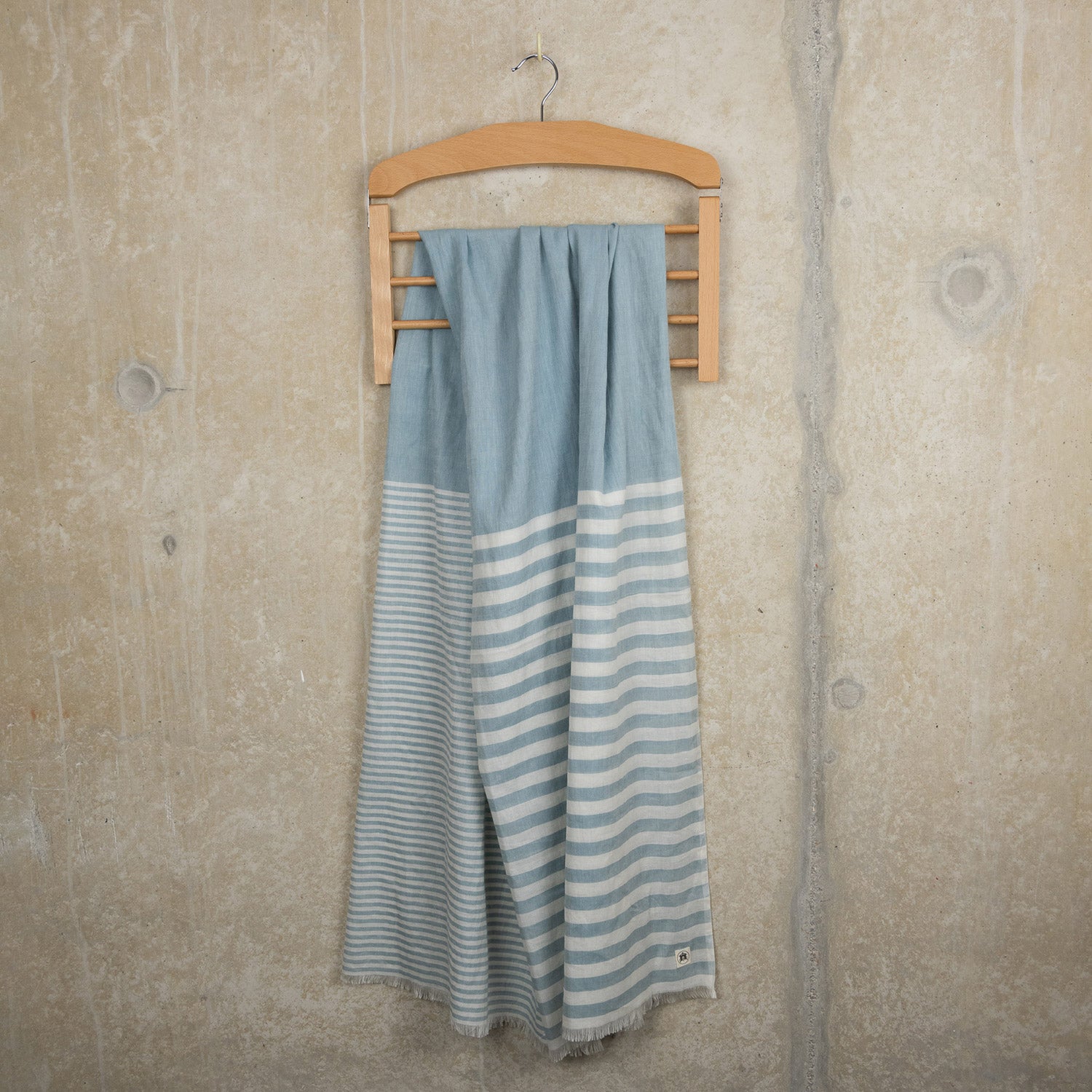 A photo of the sea blue stripe scarf on a hanger with a rustic concrete background. The scarf has blue and white stripes.